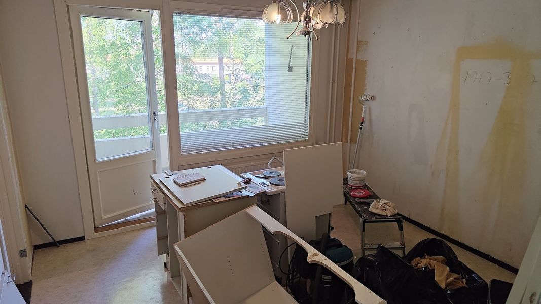 Kirkas, osittain remontoitu huone, jossa on ikkuna, parvekkeen ovi ja hajallaan olevia työkaluja ja huonekaluja.