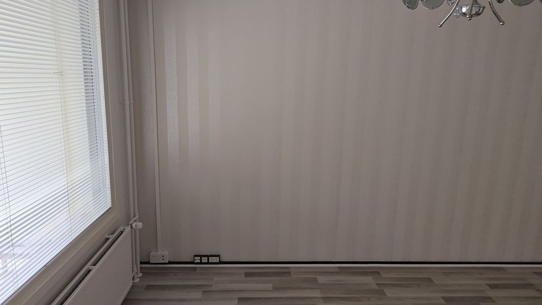 Tyhjä huoneennurkka, jossa on raidallinen tapetti, kaihtimet, patteri ja puulattia