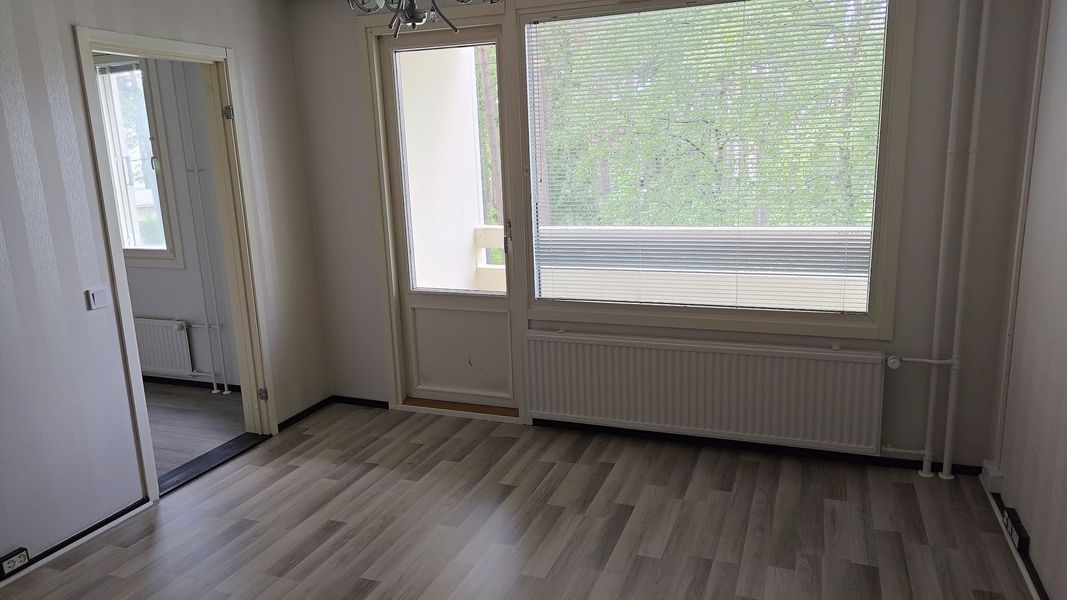 Tyhjä moderni huone, jossa on vaaleanharmaat puulattiat, valkoiset seinät ja suuret ikkunat, joiden alla on patteri.