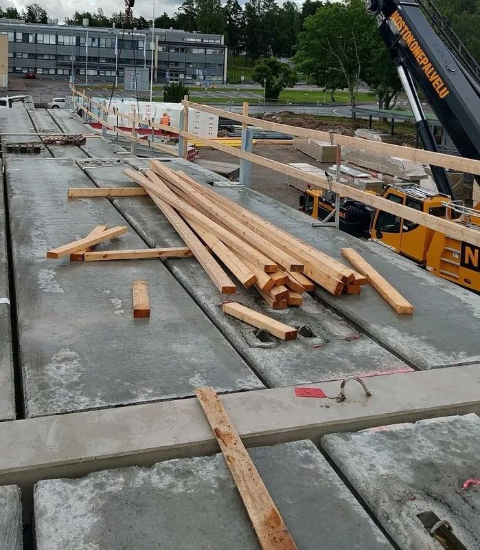 Rakennustyömaa, jossa puutavaraa on pinottu betonikatolle ja nosturinvarsi taustalla