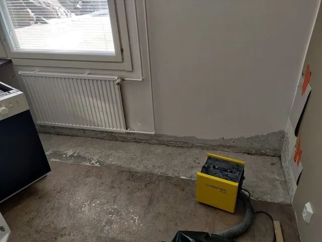 Osittain viimeistelty huone, jossa on näkyvä betonilattia, patteri ikkunan alla ja keltainen sähkötyökalu lattialla.
