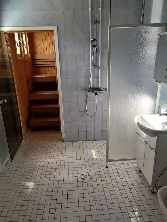Pieni laatoitettu kylpyhuone, jossa suihku, pesuallas, wc ja avoin ovi puiseen saunamaiseen huoneeseen.