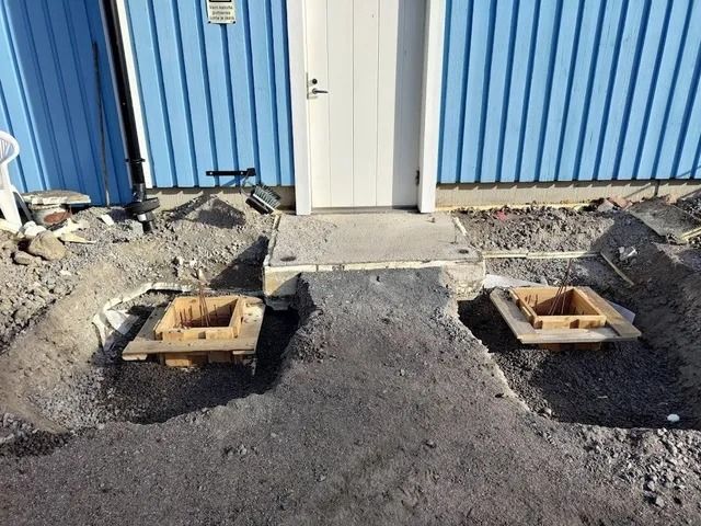 Rakennustyömaa, jossa kaksi avointa sähkölaatikkoa asfaltissa sinisen rakennuksen ja valkoisen oven vieressä
