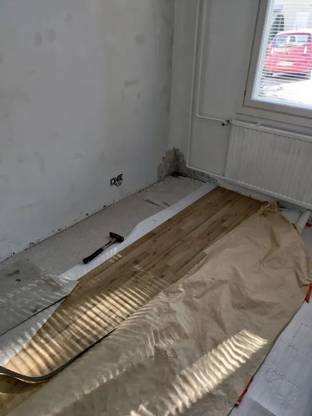 Osittain remontoitu huone, jossa on valkoiset seinät, ikkuna ja ruskea tapetti lattiaa patterin vieressä.
