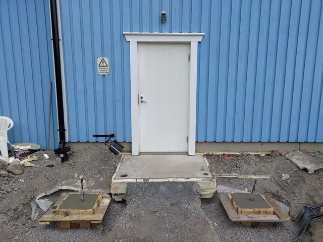 Valkoinen ovi sinisessä aaltopellissä, jossa on betoniaskelmat ja kaksi puista tasoa soralla.