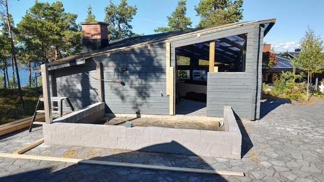 Moderni harmaa mökki, jossa on vino katto ja kivinen patio aurinkoisessa metsäisessä ympäristössä