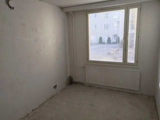 Tyhjä valkoinen huone, jossa on paljaat seinät, ikkuna, patteri ja keskeneräinen lattia.