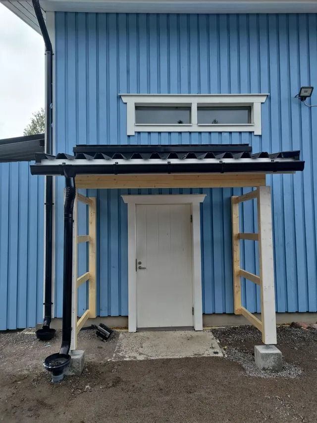Sinisen rakennuksen sisäänkäynti, jossa valkoinen ovi ja pieni katettu kuisti pylväiden varassa.