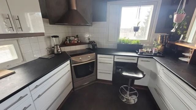 Moderni keittiö, jossa harmaat kaapistot, mustat työtasot, liesi, ikkunan vieressä oleva pesuallas ja pieni jakkara tiskillä