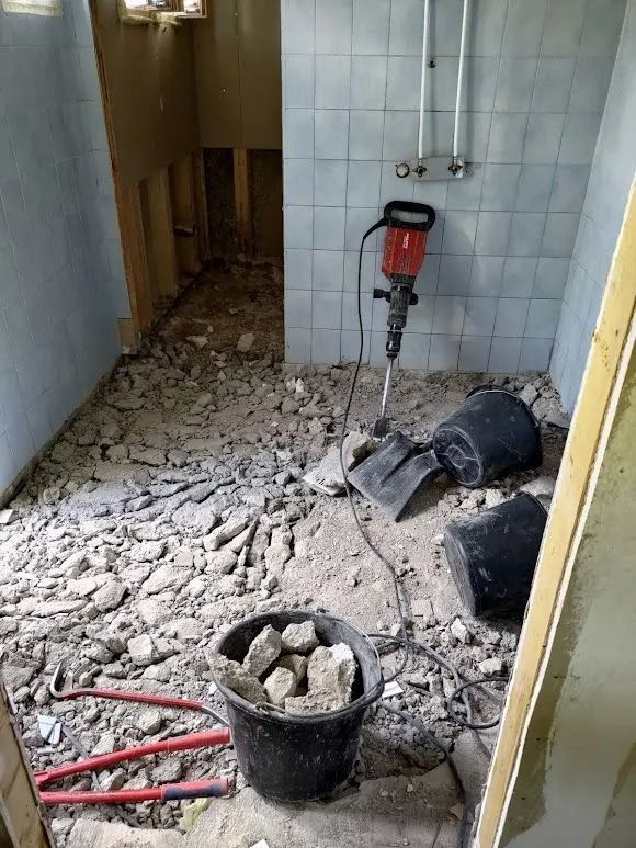 Osittain purettu laatoitettu kylpyhuone, jossa lattialla on raunioita, poravasara, ämpäreitä ja työkaluja.