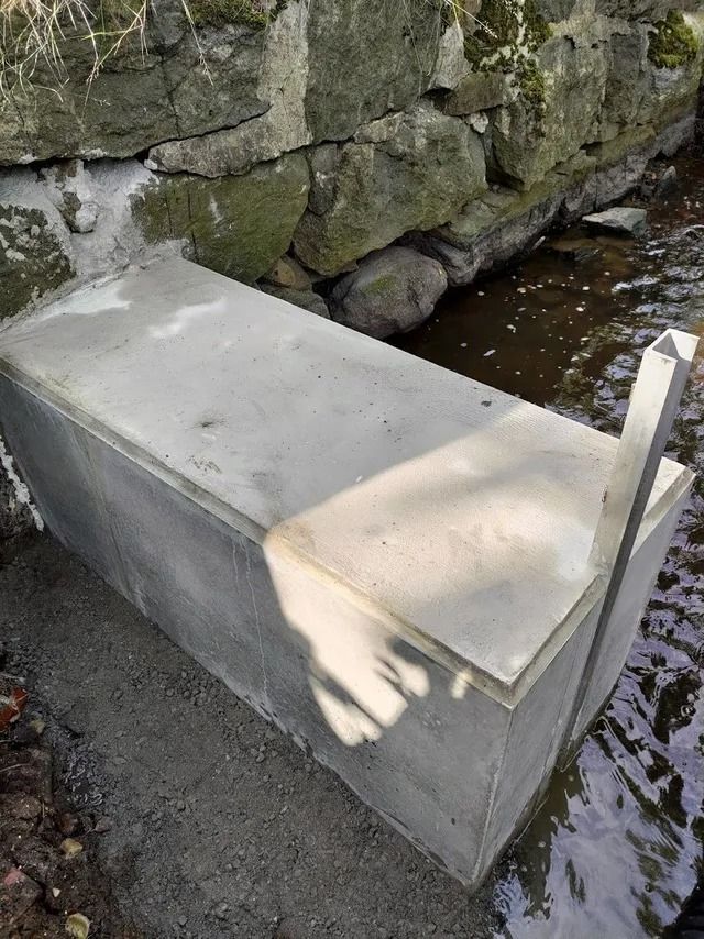 Betoninen penkki kivimuurin vieressä matalan puron varrella, auringonvalo ja varjot istuimella