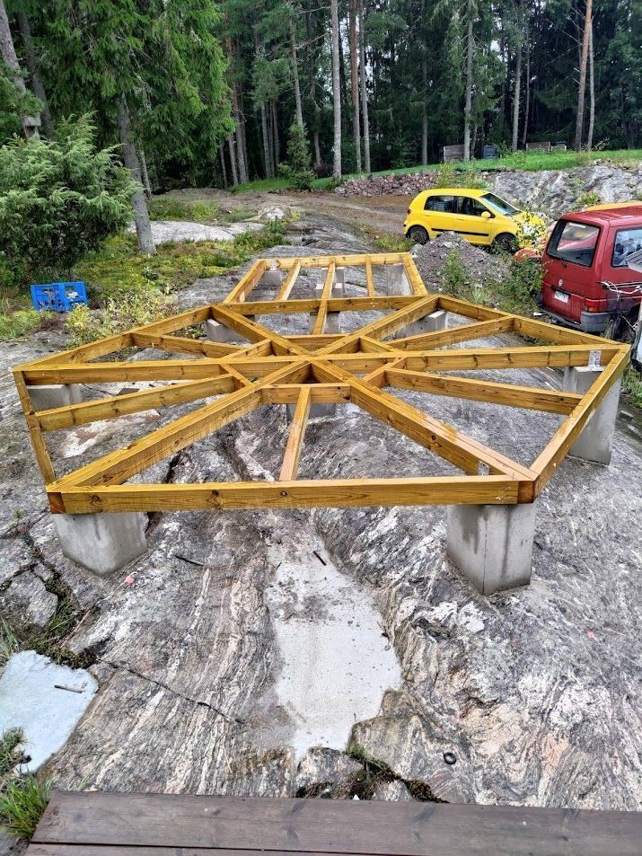 Keltainen metallirunko betonitukilla metsätyömaalla, lähellä keltainen auto ja punainen ajoneuvo