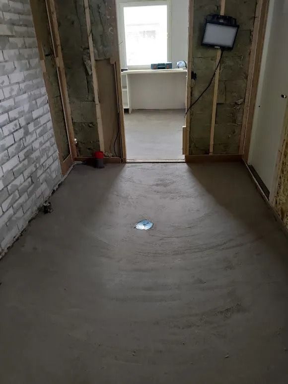 Tyhjä keskeneräinen huone, jossa on paljas betonilattia, näkyvät puupalkit ja ikkunallinen oviaukko toiseen huoneeseen.