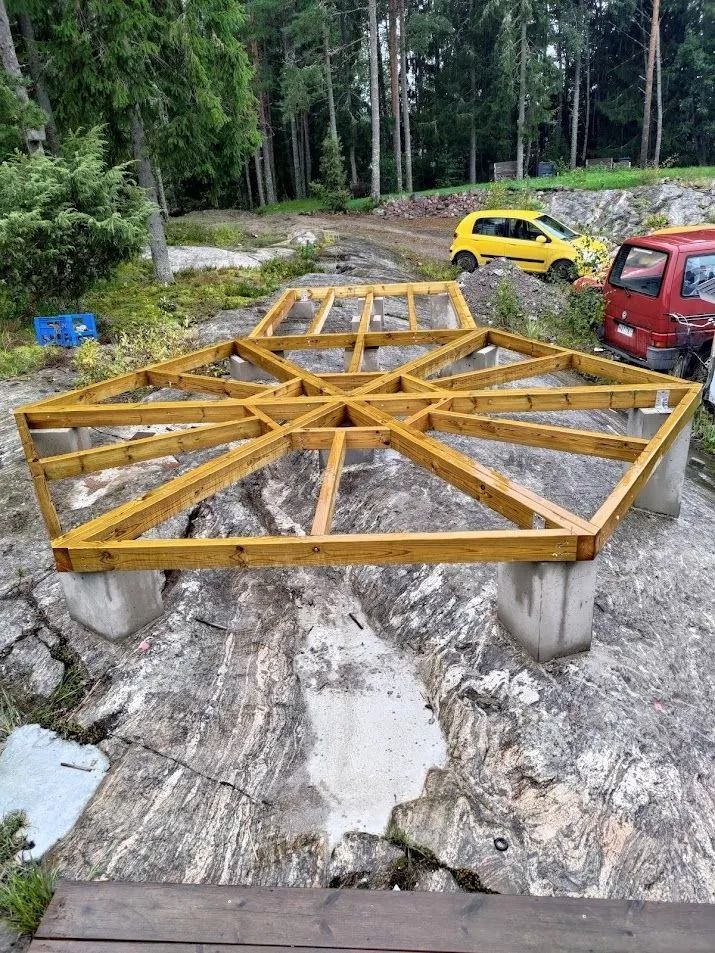 Keltainen metallirakennelma betonilohkojen päällä mutaisella hiekkatiellä, jonka vieressä autoja pysäköitynä metsään.