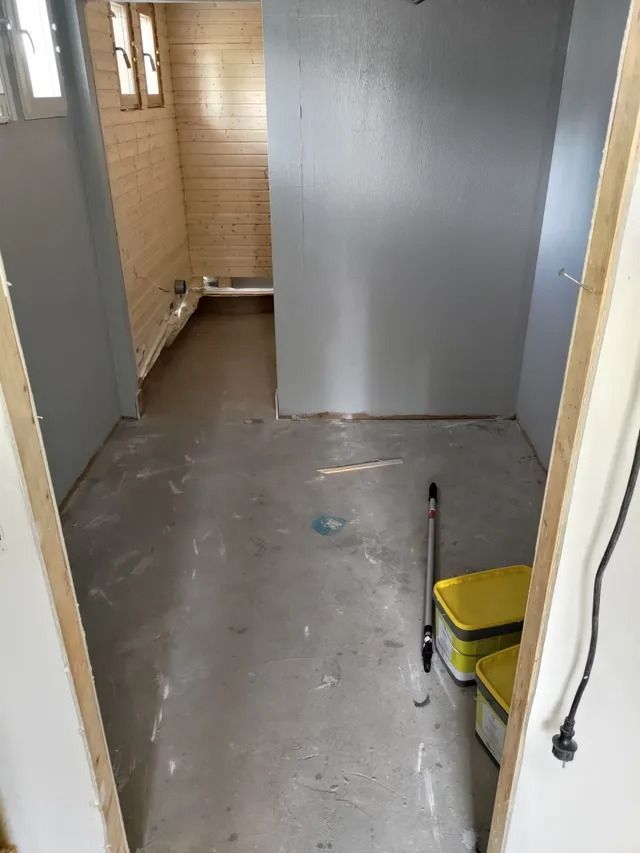 Kapea, keskeneräinen käytävä harmailla lattioilla ja seinillä, joka johtaa beigenväriseen laatoitettuun kylpyhuoneeseen.