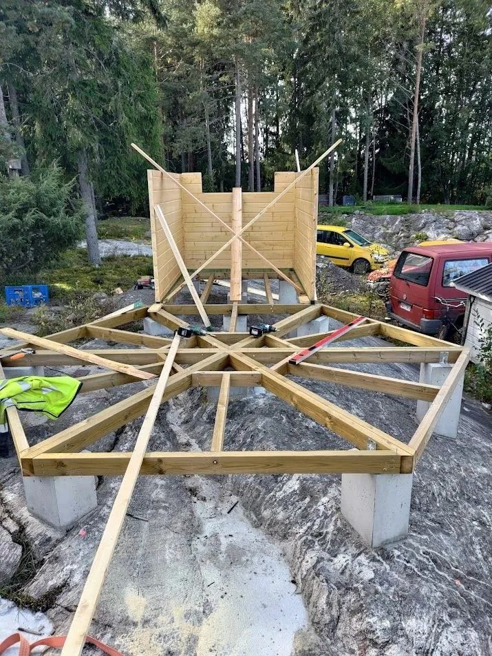 Puinen tasanne rakenteilla betonipilareilla metsäisellä tontilla, ja lähistöllä on ajoneuvoja.