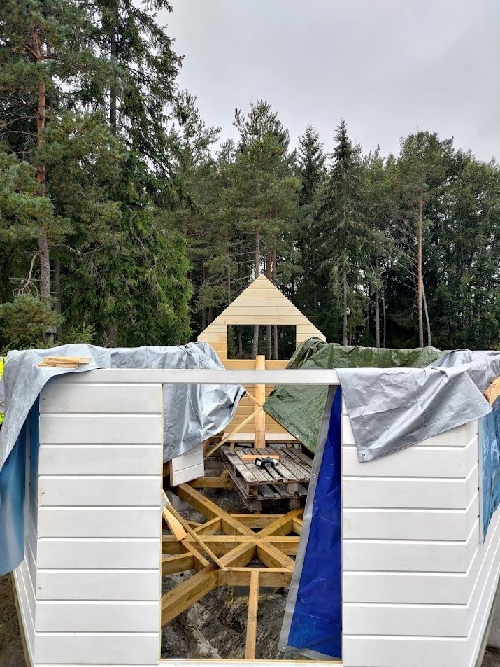 Puurunkoinen ja valkoisilla ulkoverhouspaneeleilla varustettu ulkorakennelma metsäalueella, osittain pressuilla peitettynä