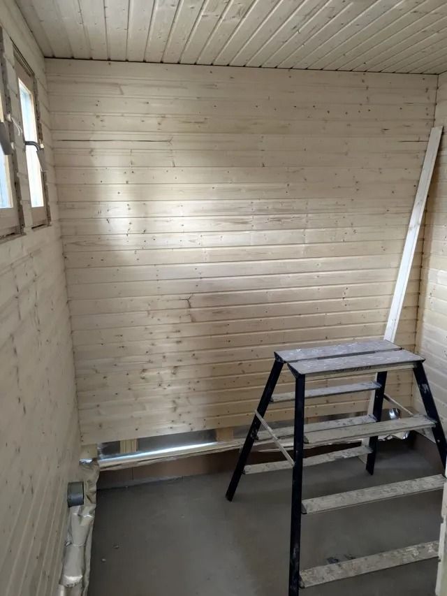 Pieni keskeneräinen huone, jossa on puupaneloidut seinät, ikkuna ja askeltikkaat betonilattialla.