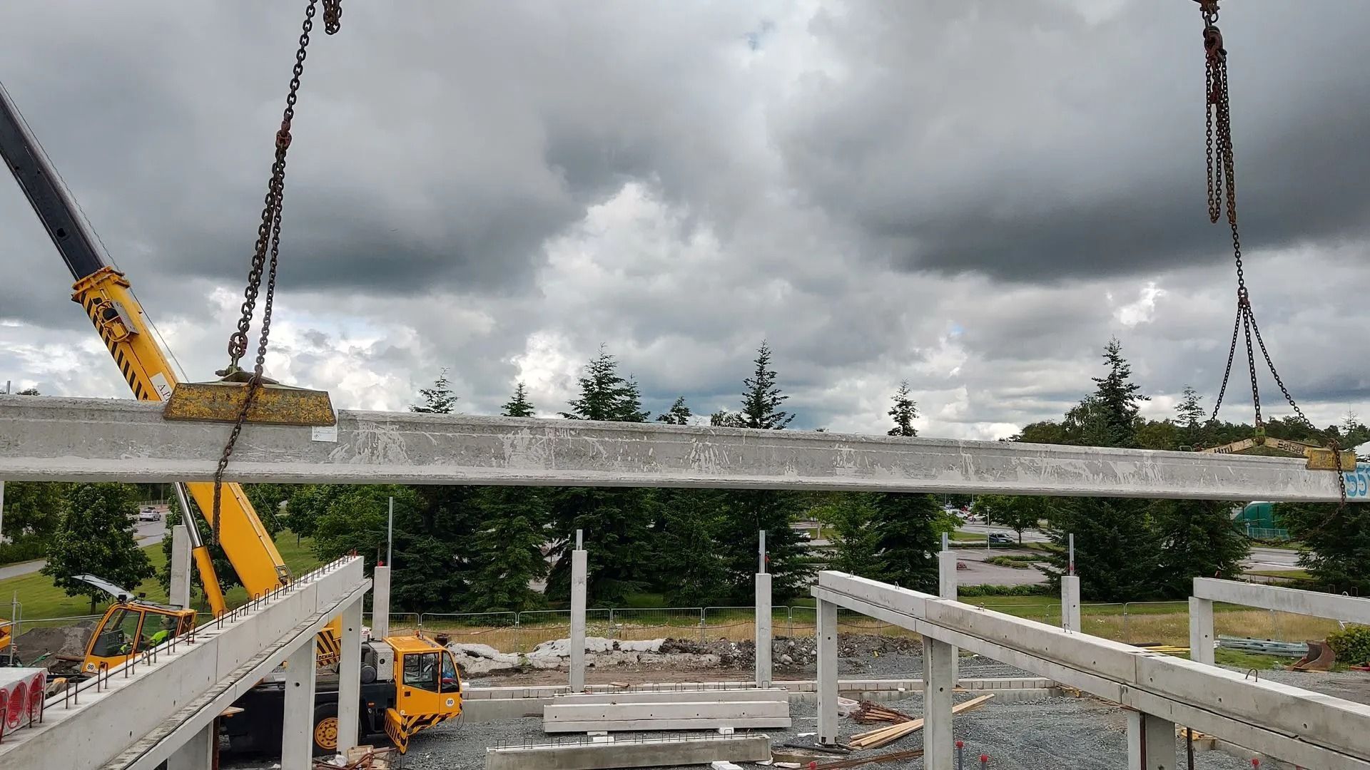 Nosturi nostaa pitkää betonipalkkia rakennustyömaan yllä pilvisen taivaan alla