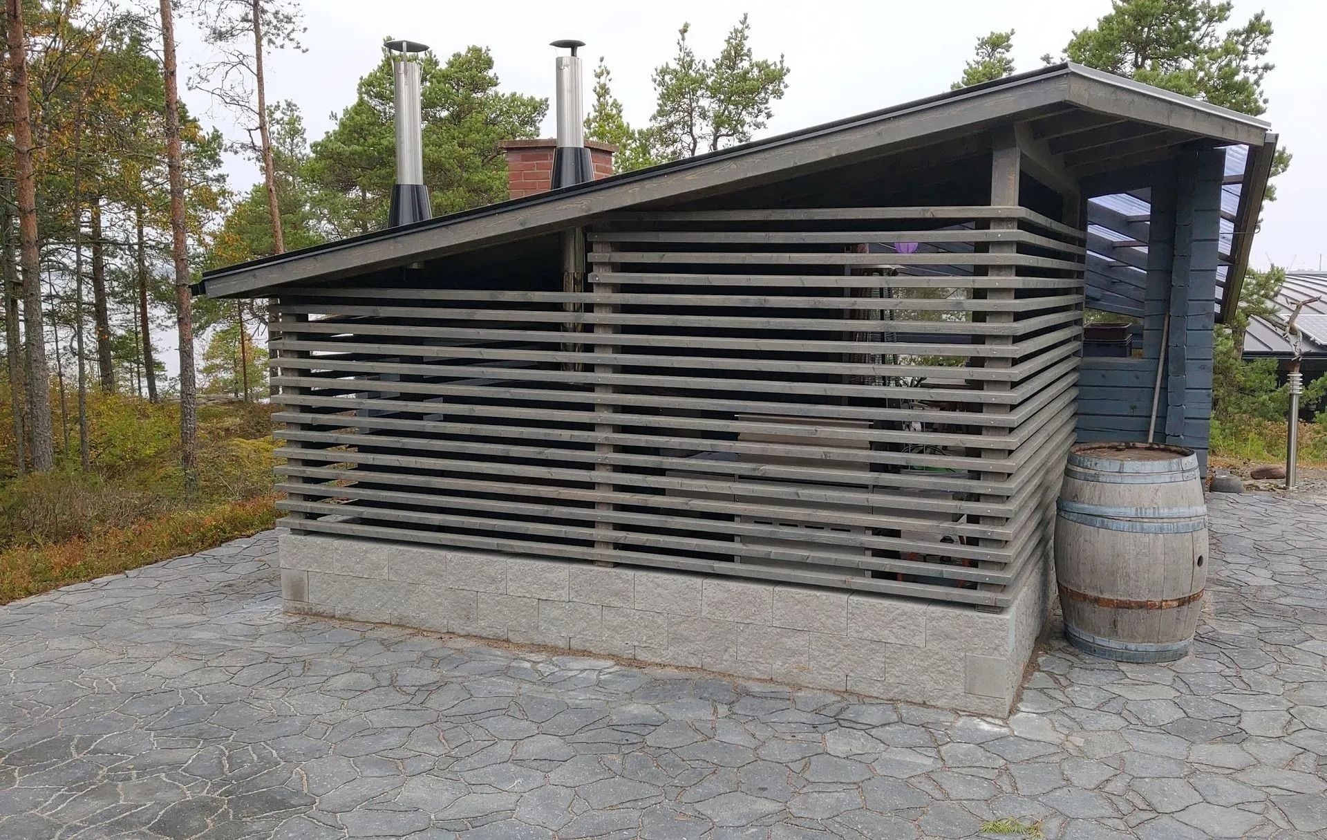 Moderni puinen mökki, jossa on vaakasuoria säleitä ja peltikatto, soratontilla, puiden ympäröimänä
