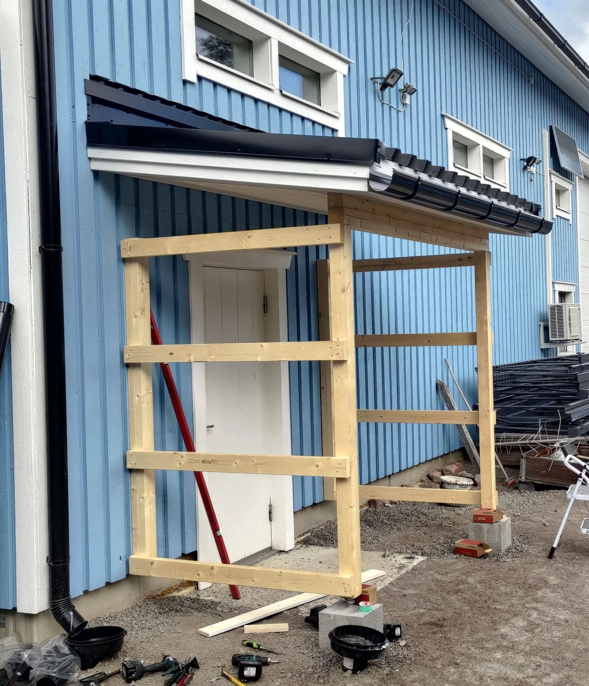 Puinen kuistin runko rakenteilla sinistä rakennusta vasten, työkaluja ja materiaaleja soralla.