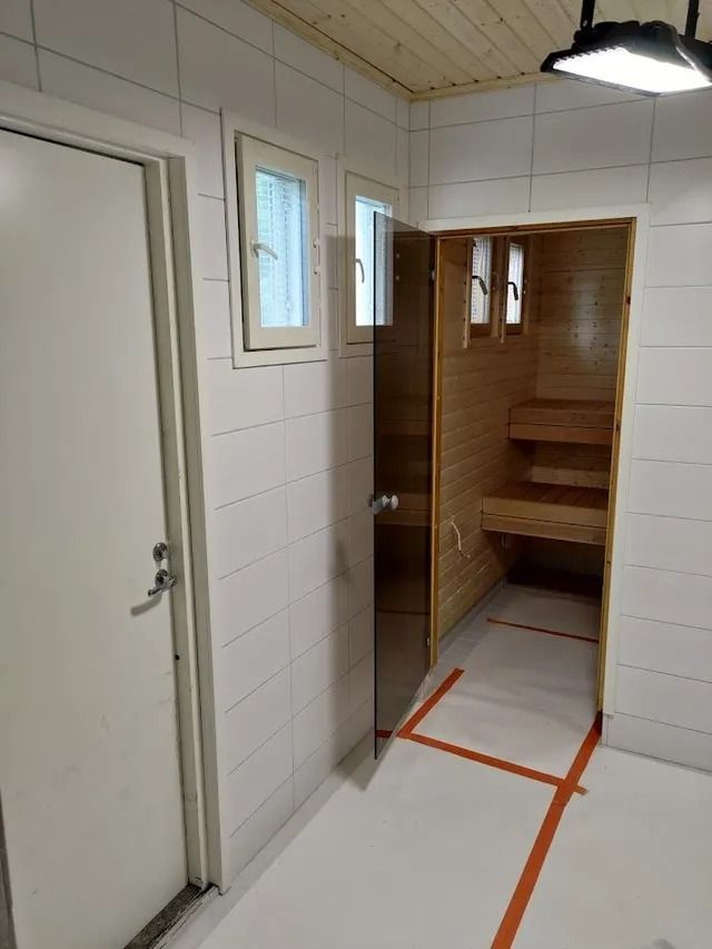 Valkoiseksi laatoitettu huone, jossa on avoin puinen saunan ovi, hyllyt sisällä, pienet ikkunat ja oranssia teippiä lattiassa.