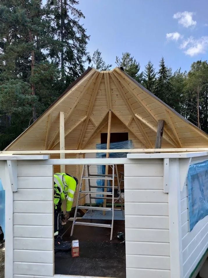 Osittain rakennettu puinen kattokehys valkoisen vajan päällä, sisällä työntekijöitä ja tikkaat