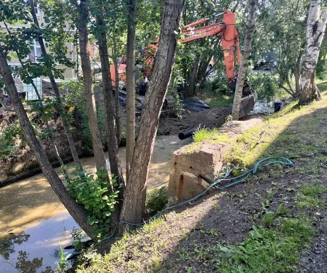 Rakennuskaivinkone työskentelee metsäisellä puronvarsialueella hiekkapolun ja betonilohkon vieressä