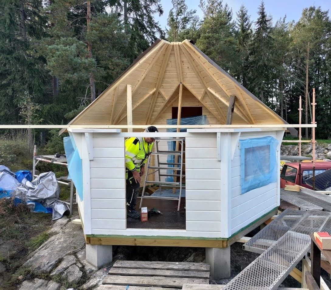Pieni kuusikulmainen mökki rakenteilla, jossa on valkoiset seinät, siniset pressuikkunat ja sisällä työntekijä.