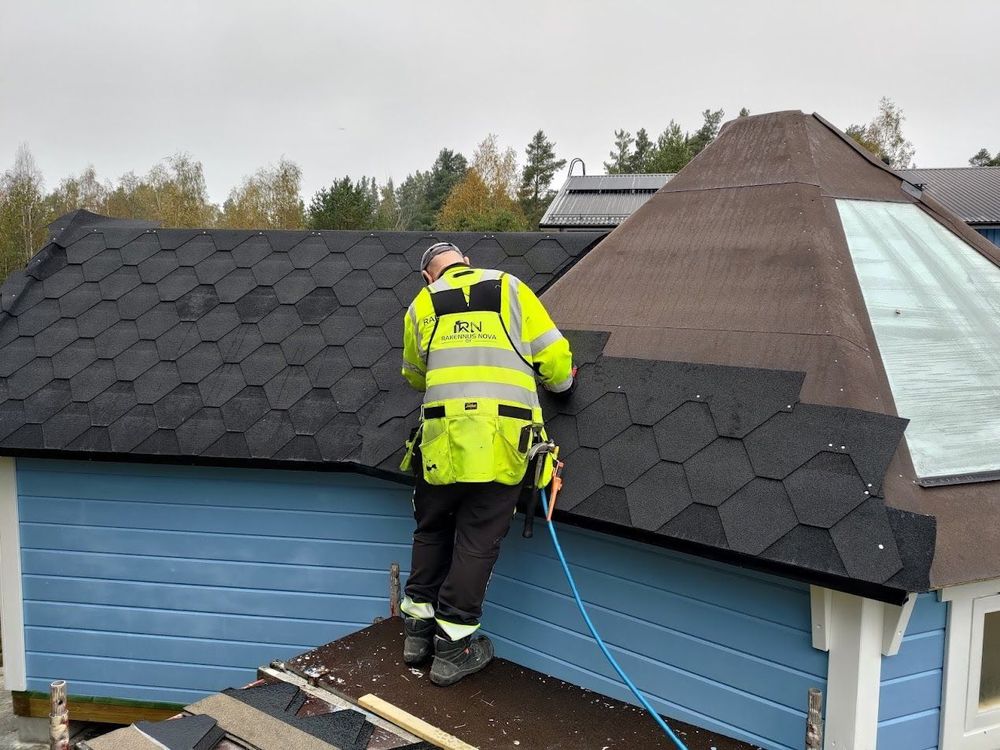 Heijastinvarusteisiin pukeutunut työntekijä korjaa tummia kattopaanuja sinisellä katolla savupiipun vieressä.