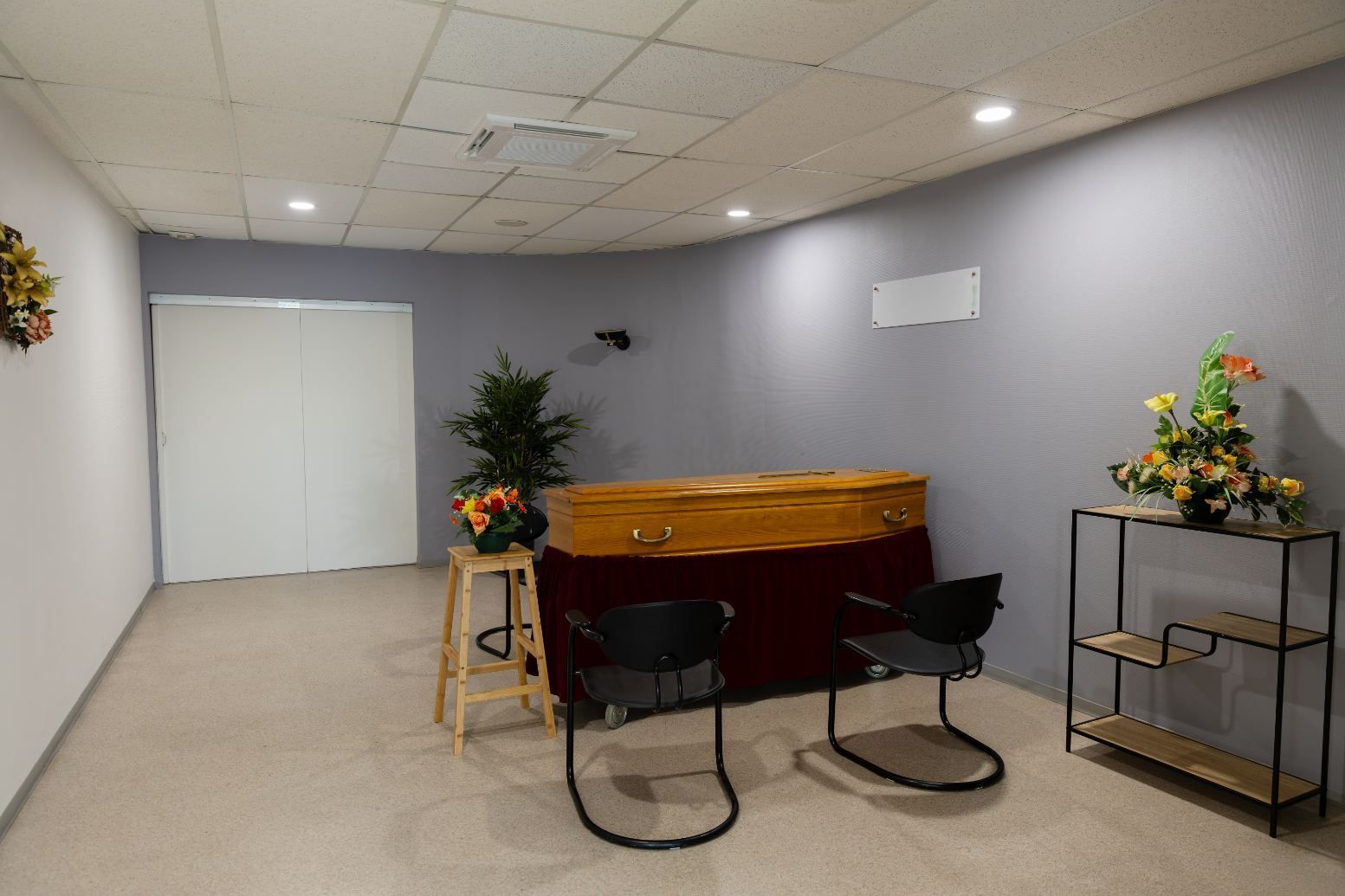 Petit espace d'attente avec une table rouge, deux chaises noires, des plantes et une étagère contre un mur gris.