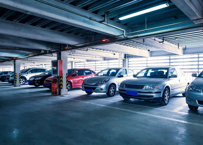 Voitures stationnées dans un parking à plusieurs niveaux. Lumières fluorescentes et poutres de soutien visibles au-dessus.
