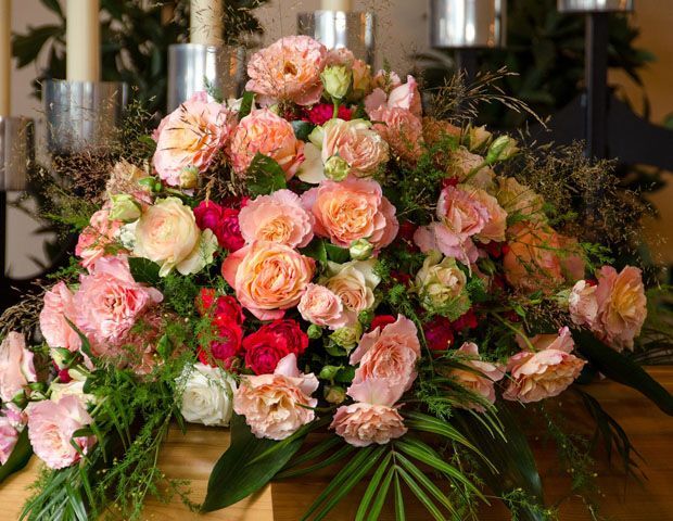 Composition florale de roses pêche et roses, d'œillets rouges, de verdure, avec des bougies en arrière-plan.