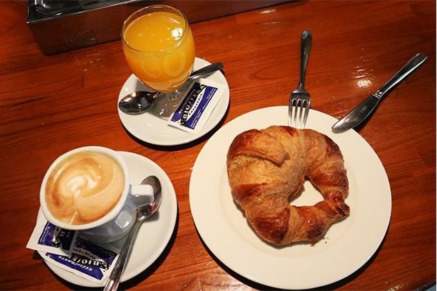 Hay un croissant en un plato junto a una taza de café y un vaso de jugo de naranja.