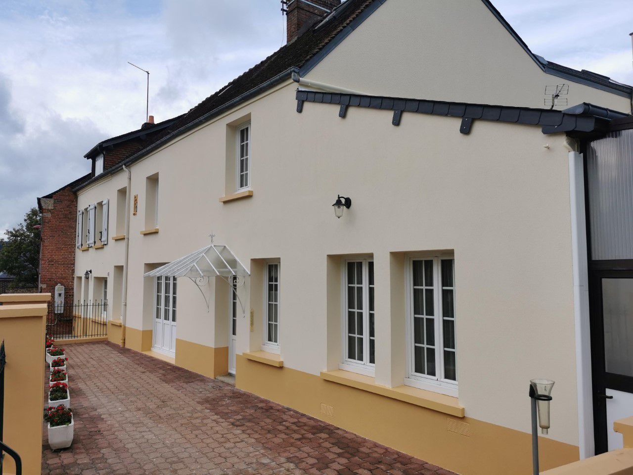 Maison de deux étages couleur crème avec des boiseries jaunes, une allée en briques et un petit auvent en verre.