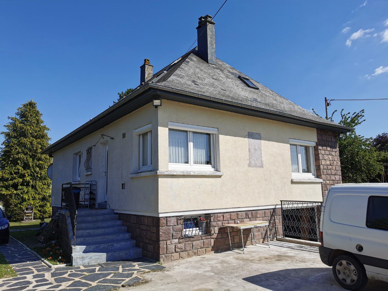 Maison en stuc beige avec façade en pierre, toit et fenêtres sombres, sur fond de ciel bleu.