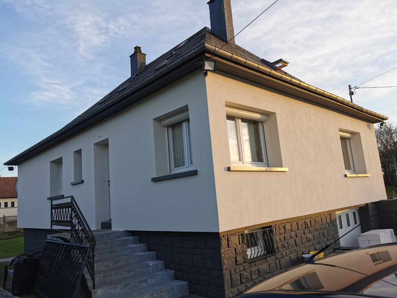 Une maison à deux étages aux murs blancs, au toit gris et aux fondations en pierre noire, baignée de lumière naturelle.