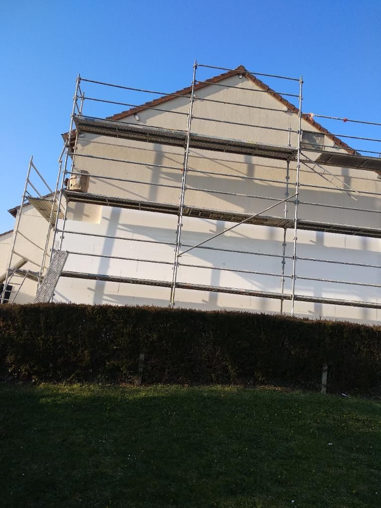 Échafaudages sur le côté d'un bâtiment en construction, murs beiges, ciel bleu, herbe verte au premier plan.