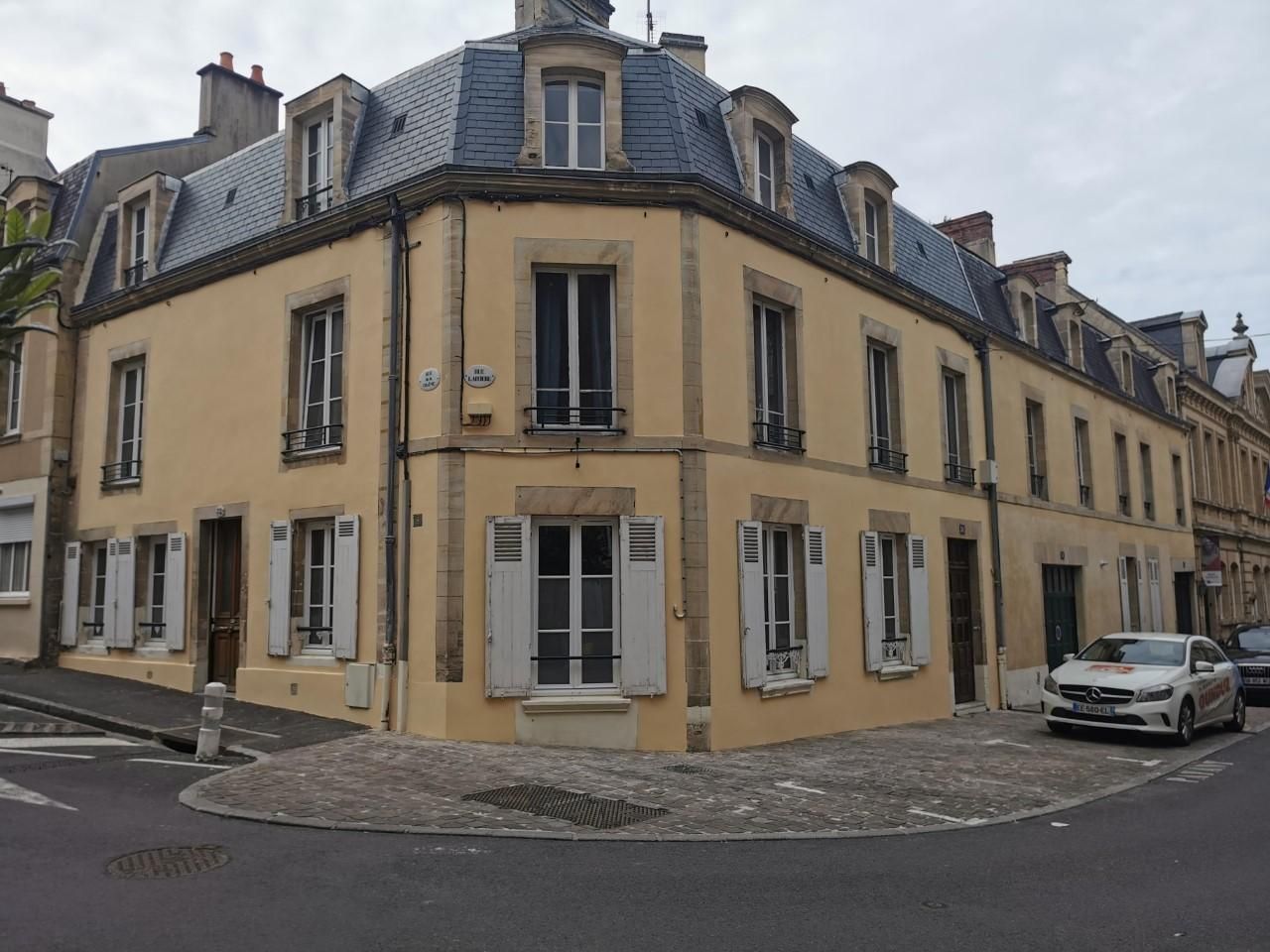 Bâtiment d'angle jaune avec toit gris, fenêtres et voiture garée dans une rue pavée.
