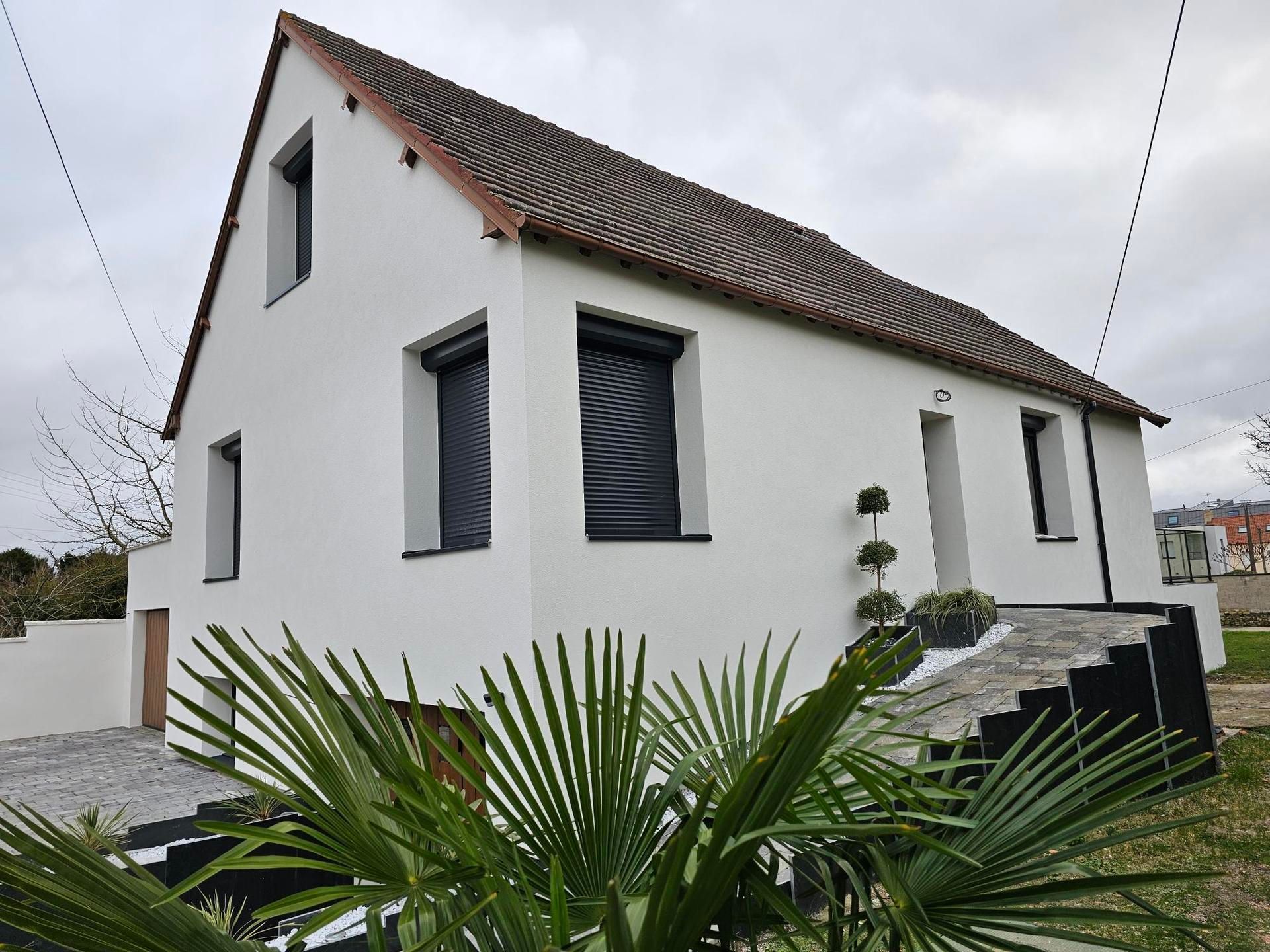Maison blanche à deux étages avec des stores sombres, des tuiles rouges et des marches menant à l'entrée ; ciel couvert.