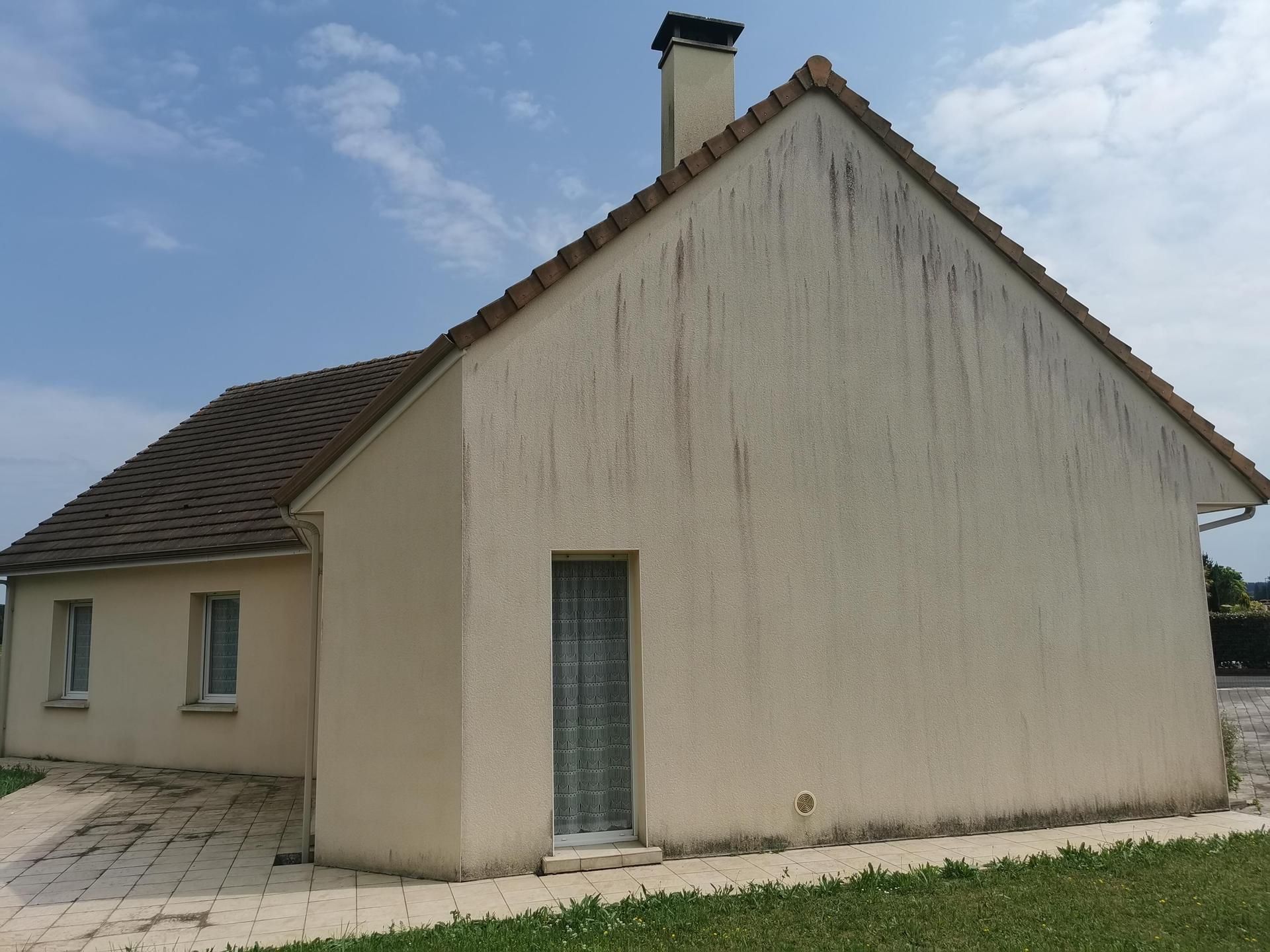Maison beige avec une toiture pentue, une cheminée et une porte vitrée étroite sous un ciel bleu