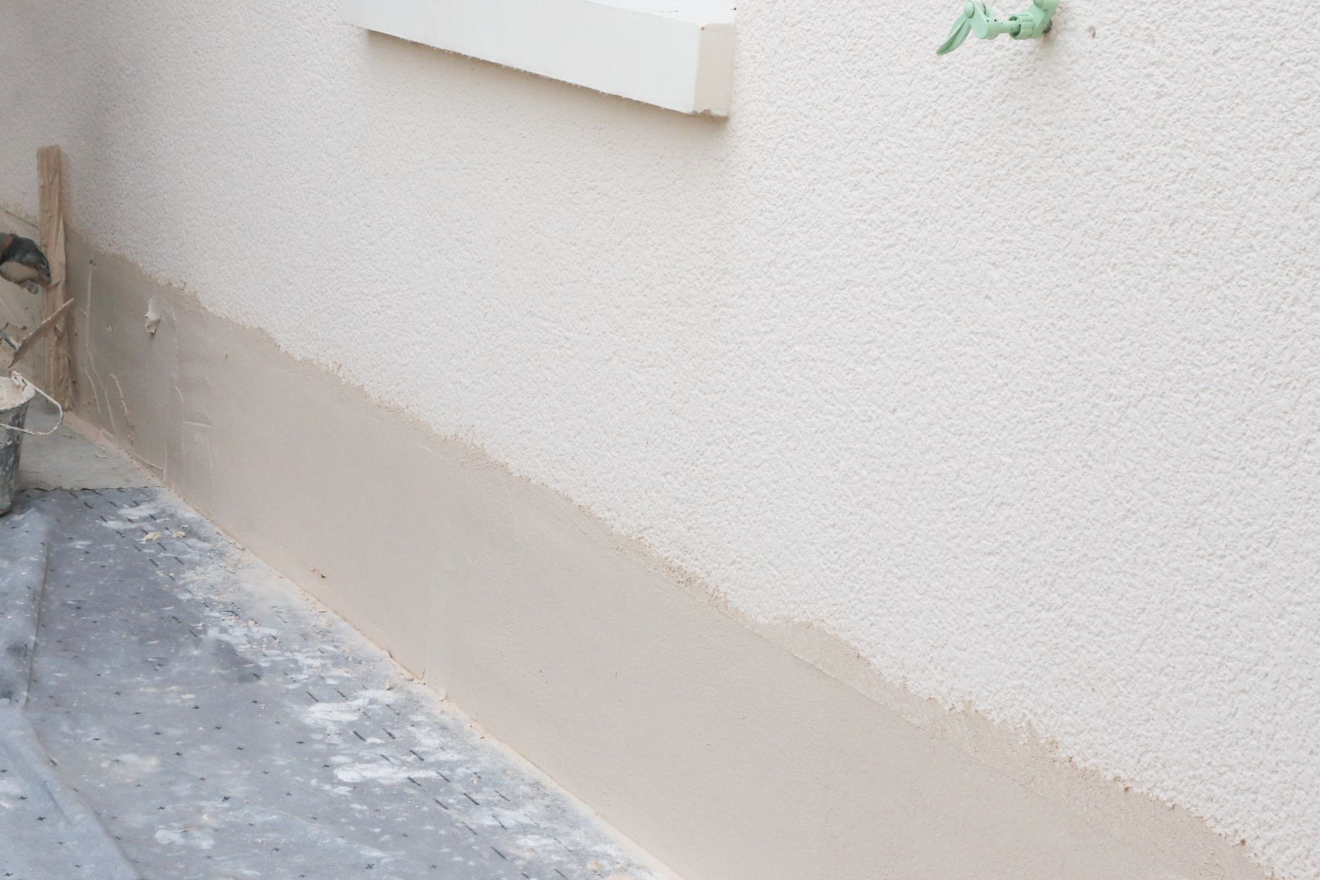 Un ouvrier applique du stuc sur la partie inférieure d'un mur extérieur ; un sol en béton est visible au premier plan.