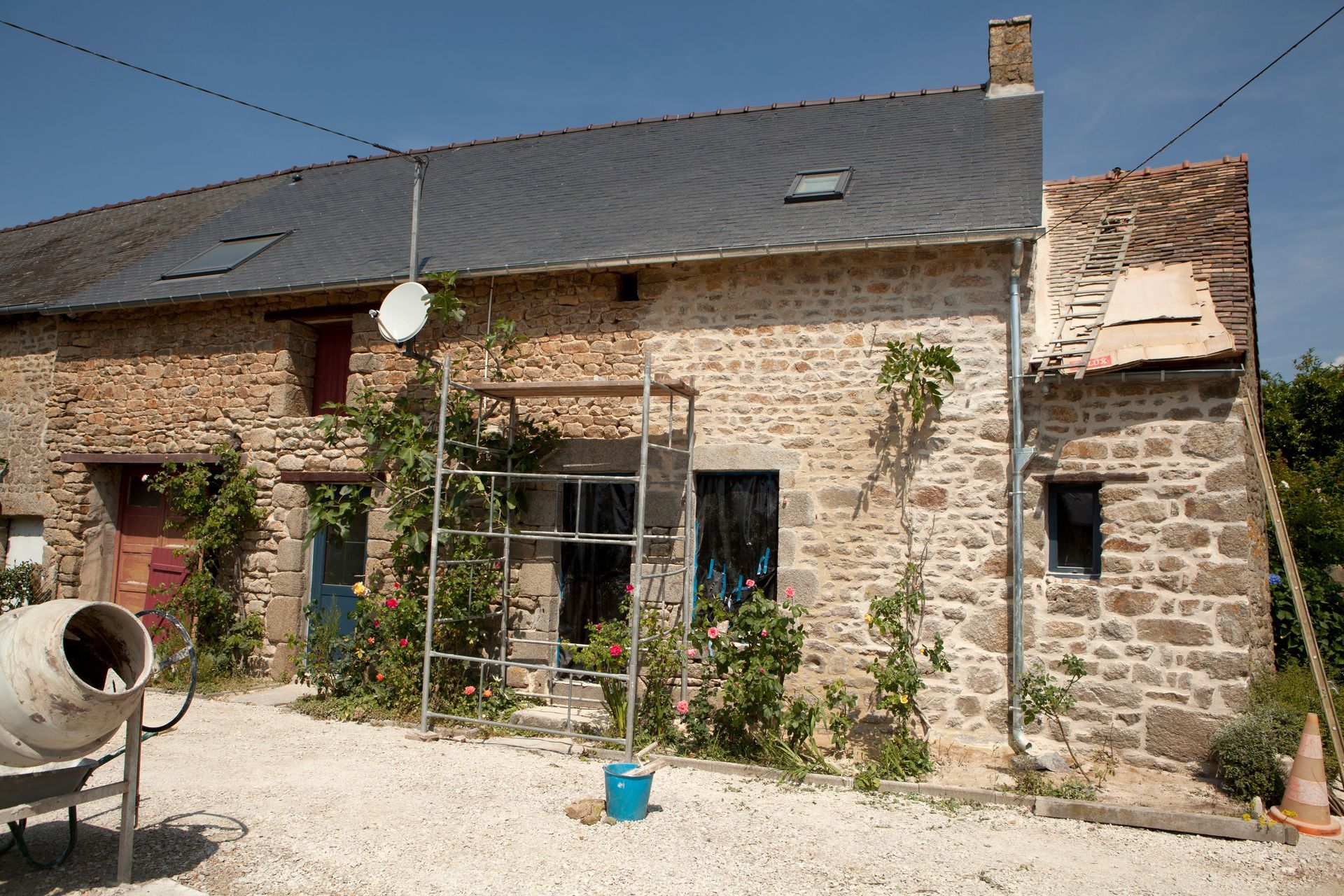 Maison en pierre à l'extérieur, échafaudages, vignes et bétonniÚre, avec un toit partiellement refait sous un ciel bleu.