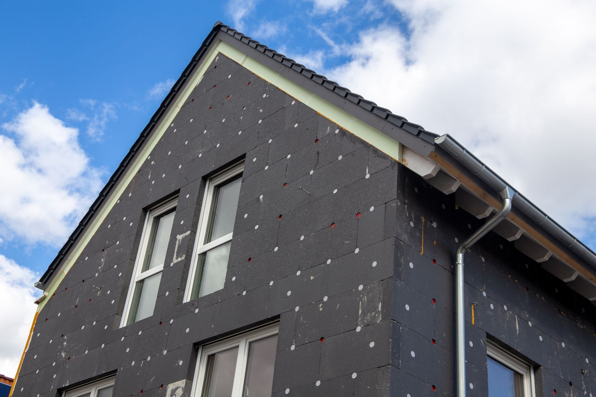 Extérieur de maison avec isolation sombre, fenêtres à cadre blanc et toit noir sur un ciel nuageux.