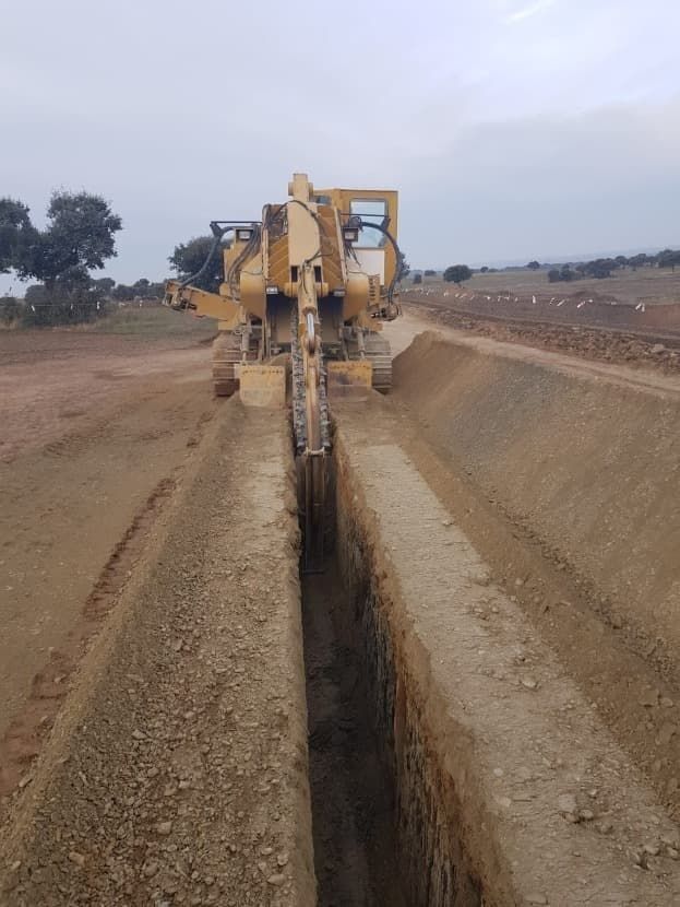 Una excavadora amarilla está cavando una zanja al costado de un camino de tierra.