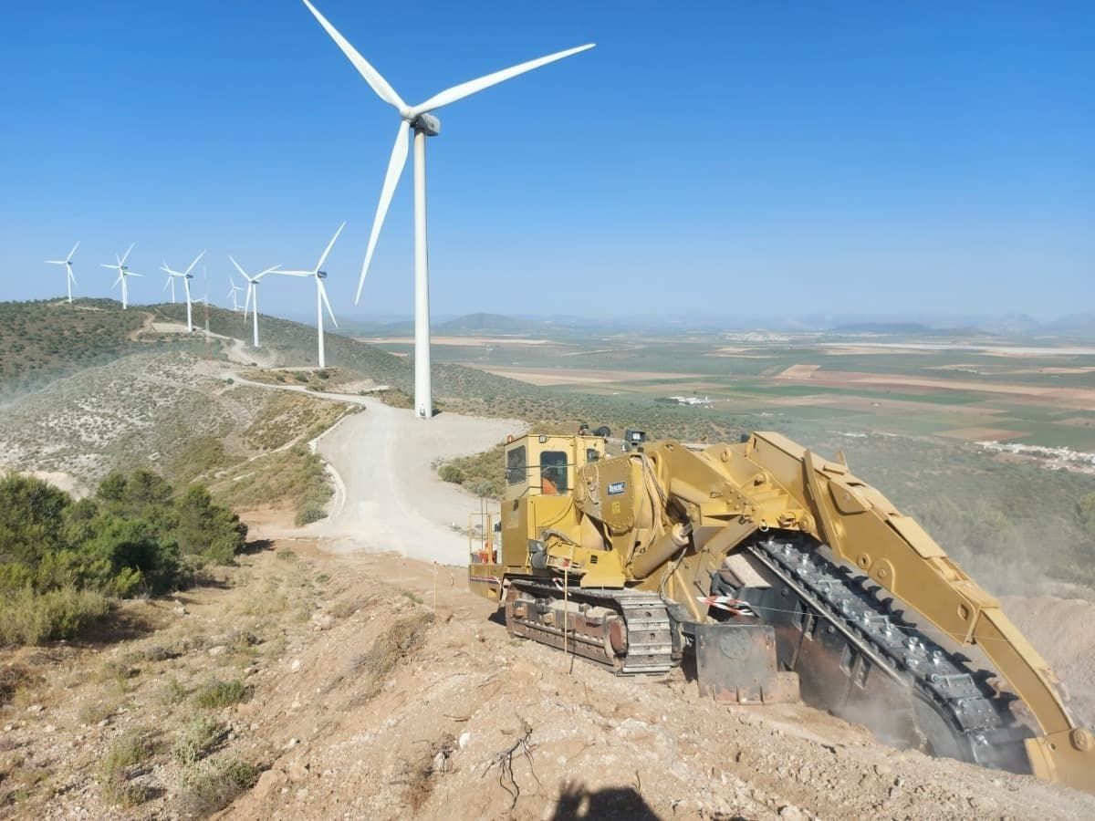 Una excavadora avanza por un camino de tierra con molinos de viento al fondo.