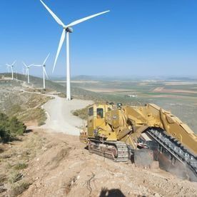 Una excavadora está trabajando en una colina al lado de turbinas eólicas.