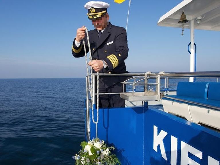 Ein Mann in Kapitänsuniform steht an der Seite eines Bootes mit der Aufschrift „Kiel“