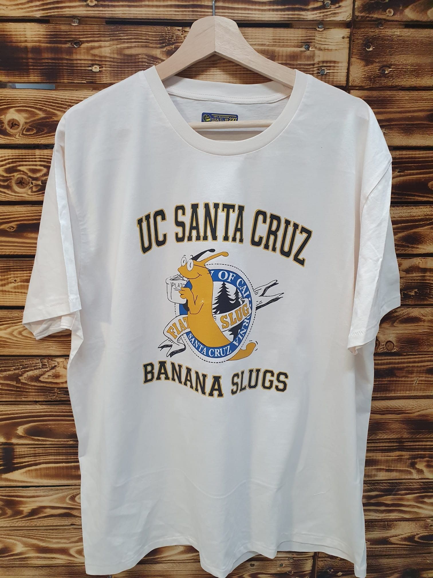 Una camiseta blanca de los Banana Slugs de la UC Santa Cruz está colgada en una percha de madera.