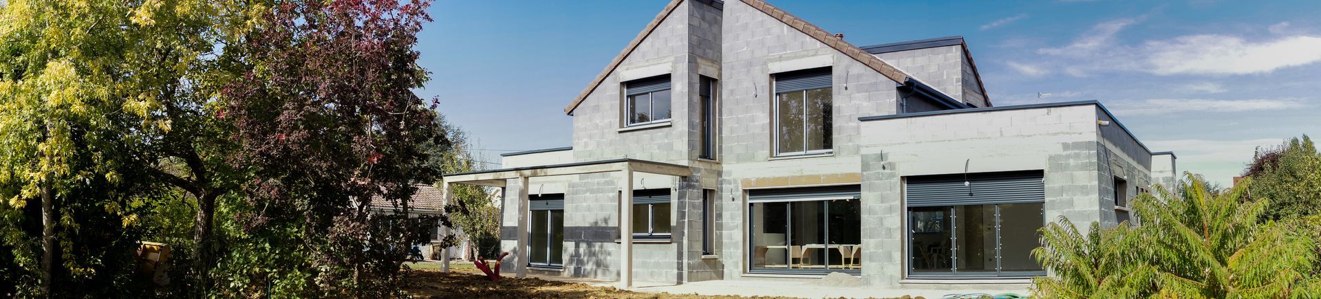 Maison à deux étages en construction, avec un toit en pente et de grandes fenêtres, entourée d'arbres.